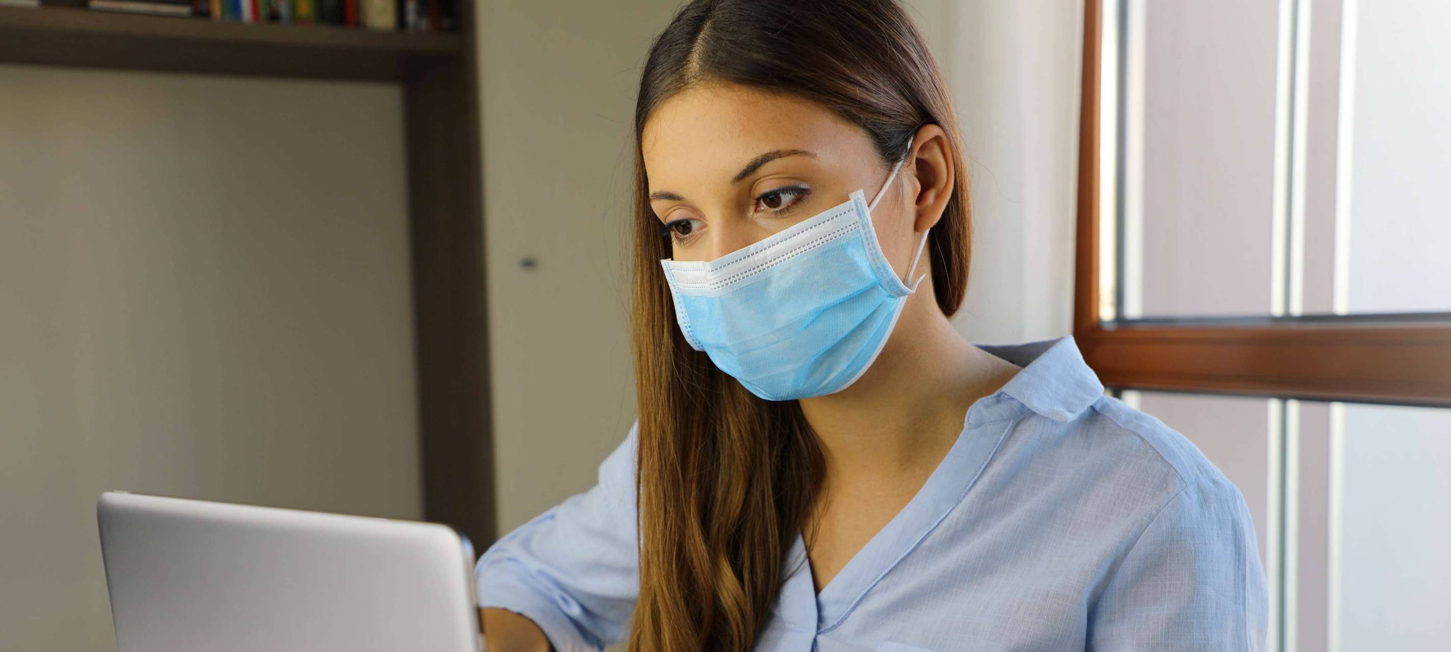 Eine Frau sitzt mit Maske vor ihrem Laptop.