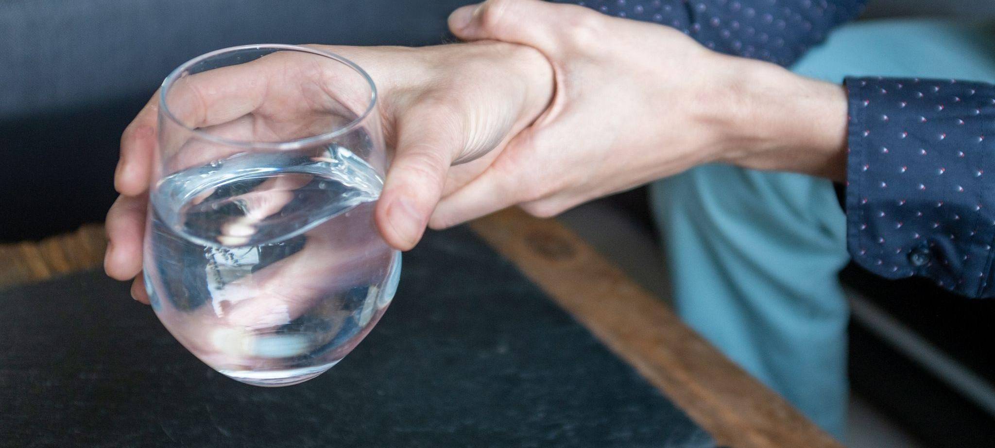 Mann hält seine zitternde Hand, die ein Wasserglas hält, fest