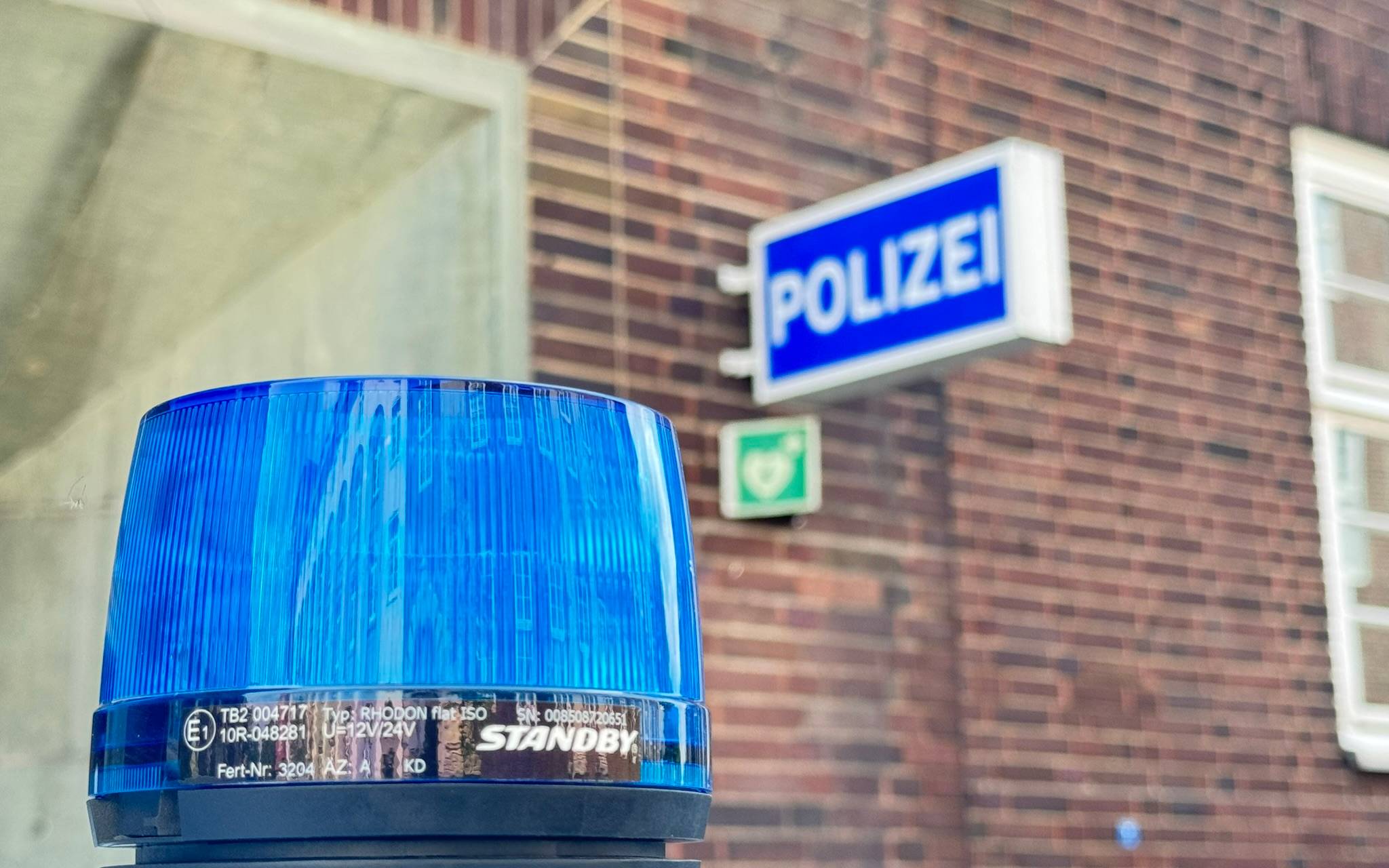 Nahaufnahme eines blauen Blaulichts (Typ Standby Rhodon) auf einem Polizeifahrzeug. Im unscharfen Hintergrund ist die Klinkerfassade einer Polizeiwache mit dem beleuchteten blauen Schild "Polizei" zu sehen.
