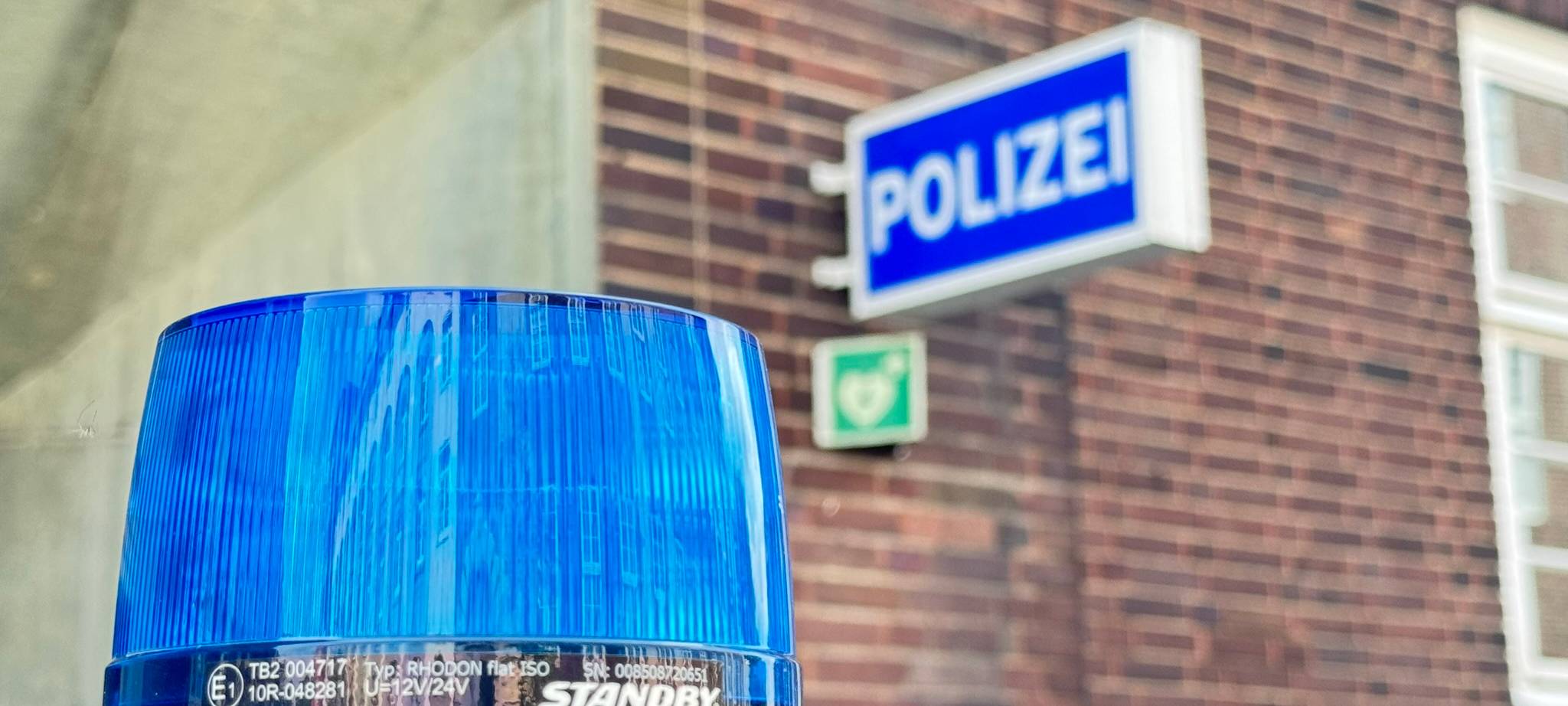 Nahaufnahme eines blauen Blaulichts (Typ Standby Rhodon) auf einem Polizeifahrzeug. Im unscharfen Hintergrund ist die Klinkerfassade einer Polizeiwache mit dem beleuchteten blauen Schild "Polizei" zu sehen.